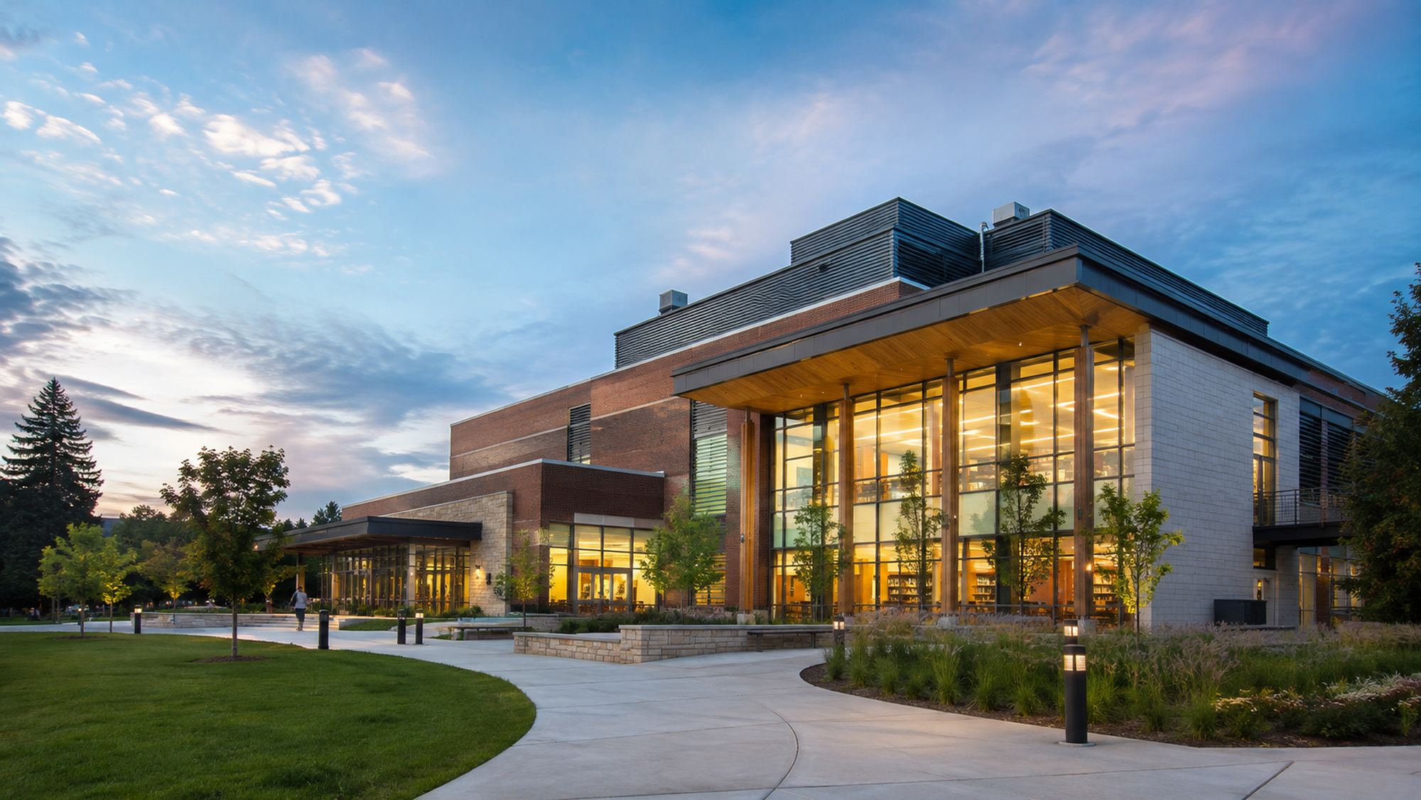 A campus building at dusk, glazed entrance hall lit from within, walkways and mature grounds in front.